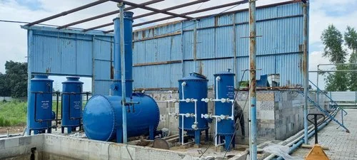 Industrial water treatment setup with multiple blue cylindrical filtration tanks, connected piping, and a horizontal pressure vessel inside a partially open metal shed.
