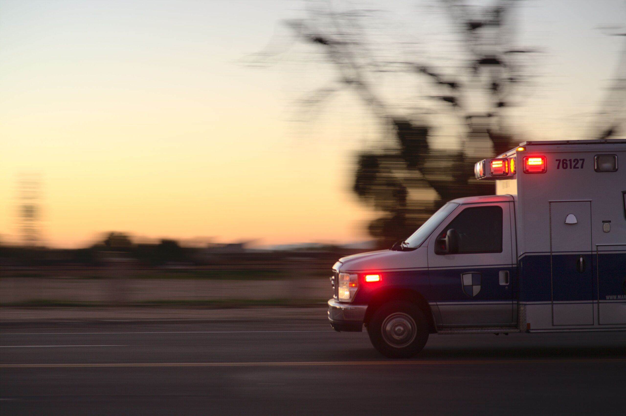 A fast-moving ambulance races down a city street at dusk, lights flashing urgently.